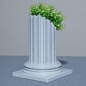 May include: A grey, fluted column planter with a square base, containing bright green foliage. The column features a classic design with vertical grooves and a rounded top. The planter is on a blue surface against a grey backdrop.