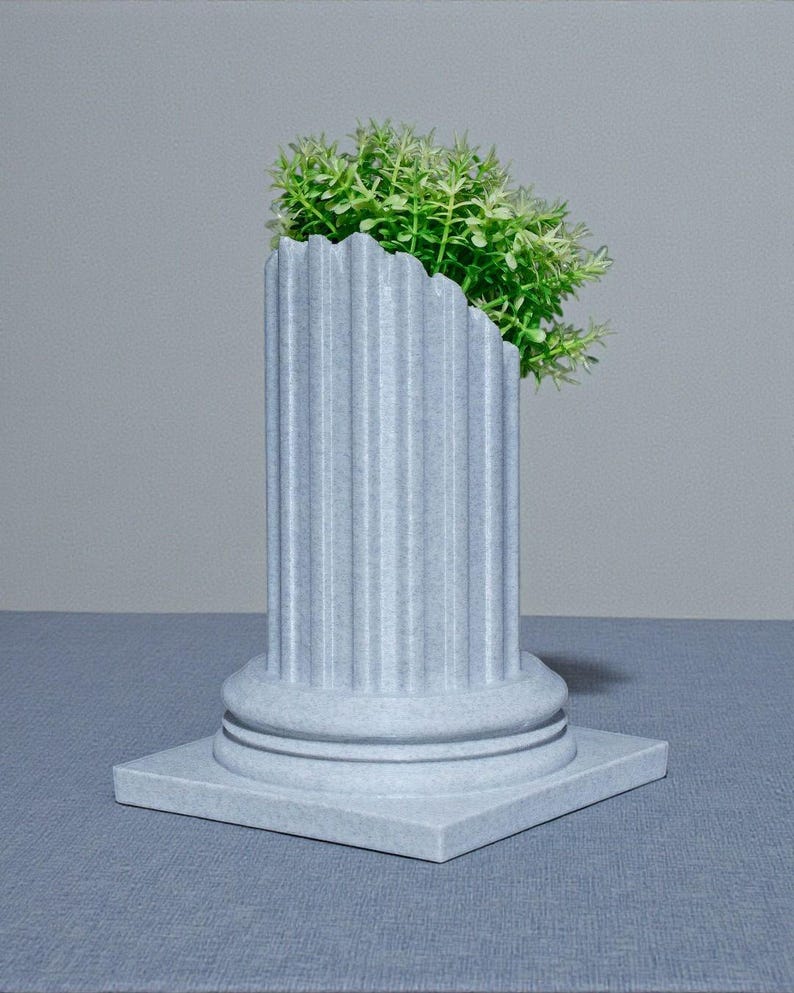 May include: A grey, fluted column planter with a square base, containing bright green foliage. The column features a classic design with vertical grooves and a rounded top. The planter is on a blue surface against a grey backdrop.