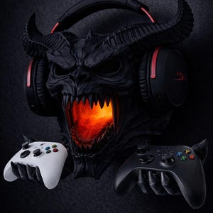 May include: A black demon-themed gaming headset stand with a glowing orange mouth. The stand holds a black and red gaming headset and two game controllers, one white and one black.