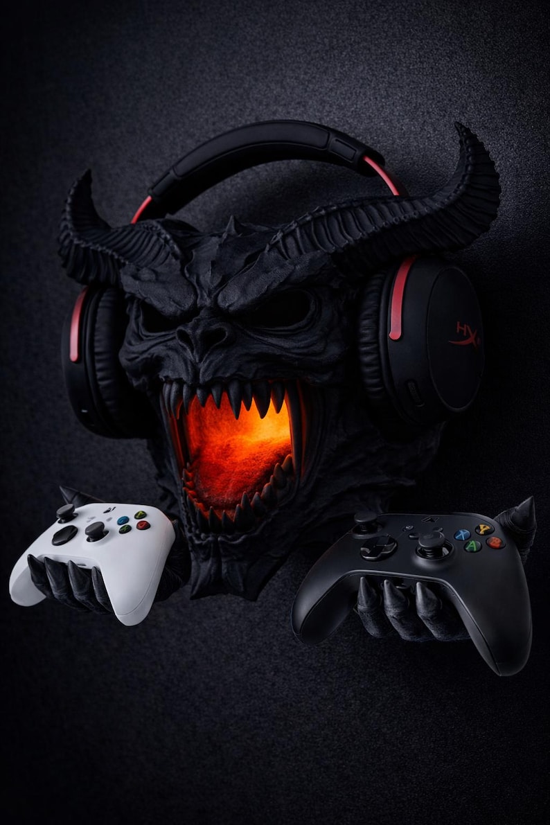 May include: A black demon-themed gaming headset stand with a glowing orange mouth. The stand holds a black and red gaming headset and two game controllers, one white and one black.