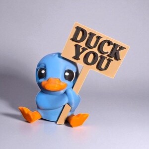 May include: A blue duck figurine with an orange beak and feet, holding a sign that reads "DUCK YOU" in black letters. The duck is sitting down, with a neutral background.