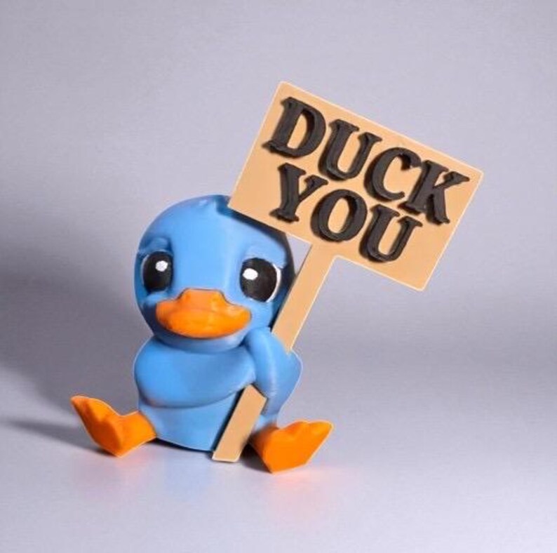 May include: A blue duck figurine with an orange beak and feet, holding a sign that reads "DUCK YOU" in black letters. The duck is sitting down, with a neutral background.