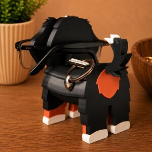 May include: A black and orange dog-shaped eyeglasses holder with a key ring. The holder has a black body with orange and white accents. A pair of eyeglasses rests on the holder. The holder is on a wooden surface.