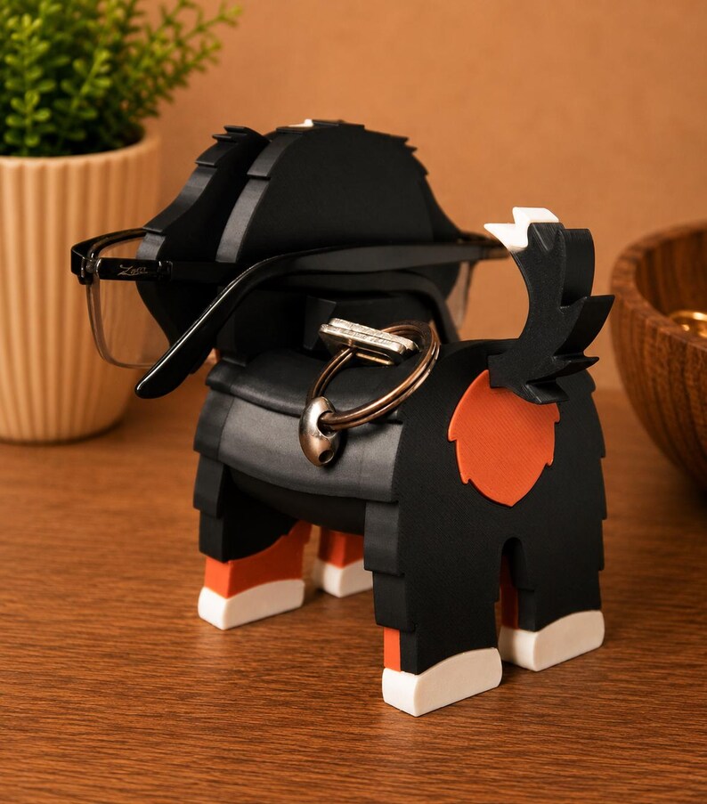 May include: A black and orange dog-shaped eyeglasses holder with a key ring. The holder has a black body with orange and white accents. A pair of eyeglasses rests on the holder. The holder is on a wooden surface.