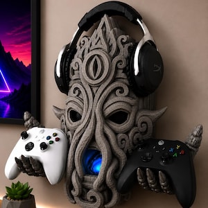 Könnte beinhalten: Ein grauer, texturierter Wandhalter in Form eines Fabelwesens mit Tentakeln, der mit zwei klauenartigen Händen einen weißen und einen schwarzen Controller hält. Ein schwarzer Over-Ear-Kopfhörer liegt oben auf und ein blaues Licht leuchtet.