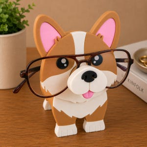 May include: A brown and white corgi-shaped eyeglass holder with pink inner ears and black eyes. The holder is wearing a pair of brown-rimmed glasses. The holder is made of a sturdy material and is designed to hold eyeglasses.