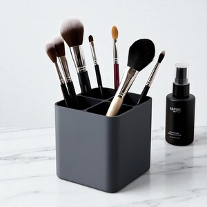 May include: A dark gray makeup brush holder with multiple compartments, filled with various makeup brushes. A black spray bottle with white text is to the right. The brushes have black handles and different brush heads.