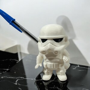 May include: A small, off-white figurine of a Stormtrooper from Star Wars. The figure has black eye lenses and is standing on a black and white surface. A blue pen is in the background.