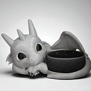 May include: A gray dragon-shaped speaker holder. The dragon has large eyes, horns, and wings, cradling a black speaker. The speaker is round and has a fabric-like texture. The dragon is resting on a white surface.