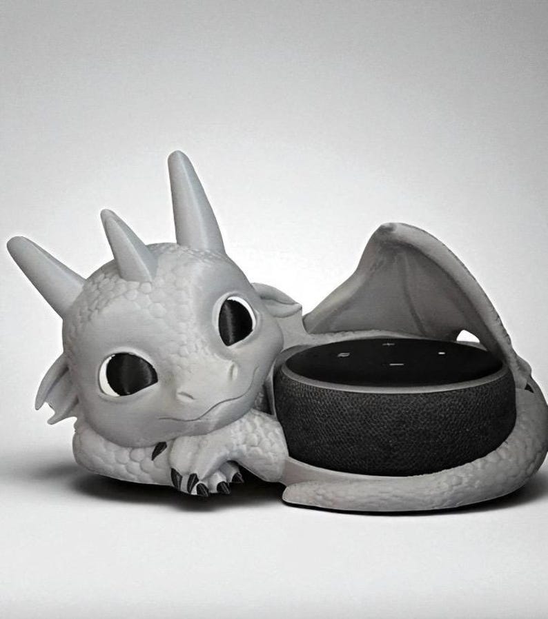 May include: A gray dragon-shaped speaker holder. The dragon has large eyes, horns, and wings, cradling a black speaker. The speaker is round and has a fabric-like texture. The dragon is resting on a white surface.