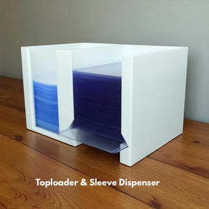 May include: A white toploader and sleeve dispenser with a clear plastic tray. The dispenser holds stacks of blue and clear plastic sleeves. The text "Toploader & Sleeve Dispenser" is printed at the bottom.