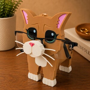 May include: A brown cat-shaped eyeglasses holder with black-framed glasses. The cat has white whiskers, a pink nose, and green eyes. The ears have pink accents, and the paws are white. The holder is on a wooden surface.