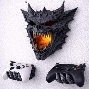 May include: A black and gray demon skull wall mount with a fiery interior, flanked by two game controller holders. The text "mounts to wall with screws (not included)" is above the skull. One controller holder is white, the other black.