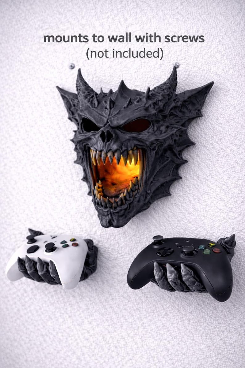 May include: A black and gray demon skull wall mount with a fiery interior, flanked by two game controller holders. The text "mounts to wall with screws (not included)" is above the skull. One controller holder is white, the other black.