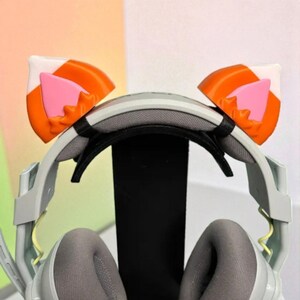 May include: Gray gaming headset with attached cat ear accessories. The cat ears are orange, white, and pink. The headset has gray earcups and a black headband. The microphone is on the left side.