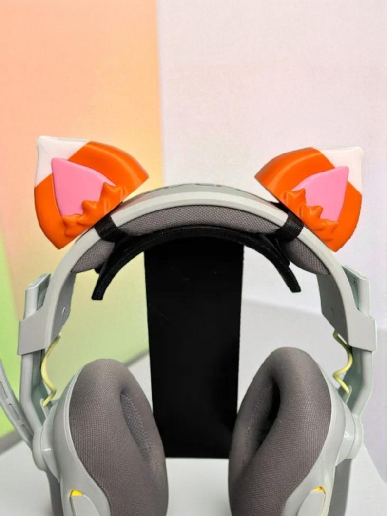 May include: Gray gaming headset with attached cat ear accessories. The cat ears are orange, white, and pink. The headset has gray earcups and a black headband. The microphone is on the left side.