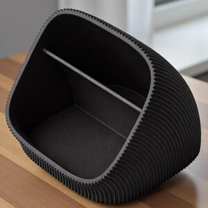 May include: A black desk tidy with a distinctive, textured design. The organiser has a curved shape with a ridged exterior and a divider inside. It sits on a wooden surface.