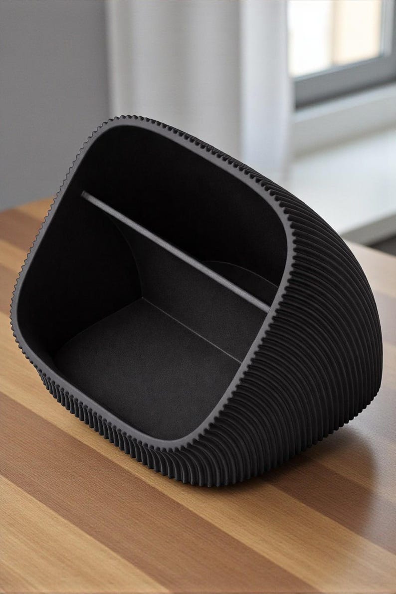 May include: A black desk tidy with a distinctive, textured design. The organiser has a curved shape with a ridged exterior and a divider inside. It sits on a wooden surface.