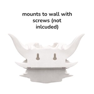 Puede incluir: Escultura decorativa blanca para montar en la pared con dise&ntilde;o de calavera con cuernos. La escultura tiene dos ranuras para colgar y el texto "mounts to wall with screws (not included)".