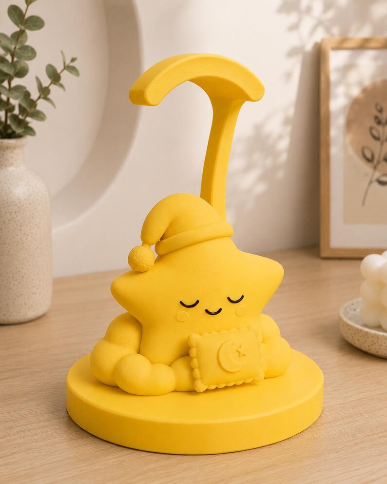 May include: A yellow star-shaped night light with a sleeping cap and a crescent moon design on a pillow. The lamp has a curved arm and sits on a round base. The lamp is made of a soft material.