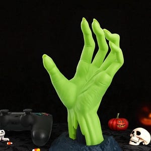 Puede incluir: Una mano verde estilizada, que emerge de una base azul oscuro, con una superficie texturizada. La mano est&aacute; posicionada como si se extendiera. Decoraciones tem&aacute;ticas de Halloween, incluyendo una calabaza y una calavera, en el fondo.