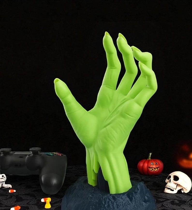 Puede incluir: Una mano verde estilizada, que emerge de una base azul oscuro, con una superficie texturizada. La mano est&aacute; posicionada como si se extendiera. Decoraciones tem&aacute;ticas de Halloween, incluyendo una calabaza y una calavera, en el fondo.