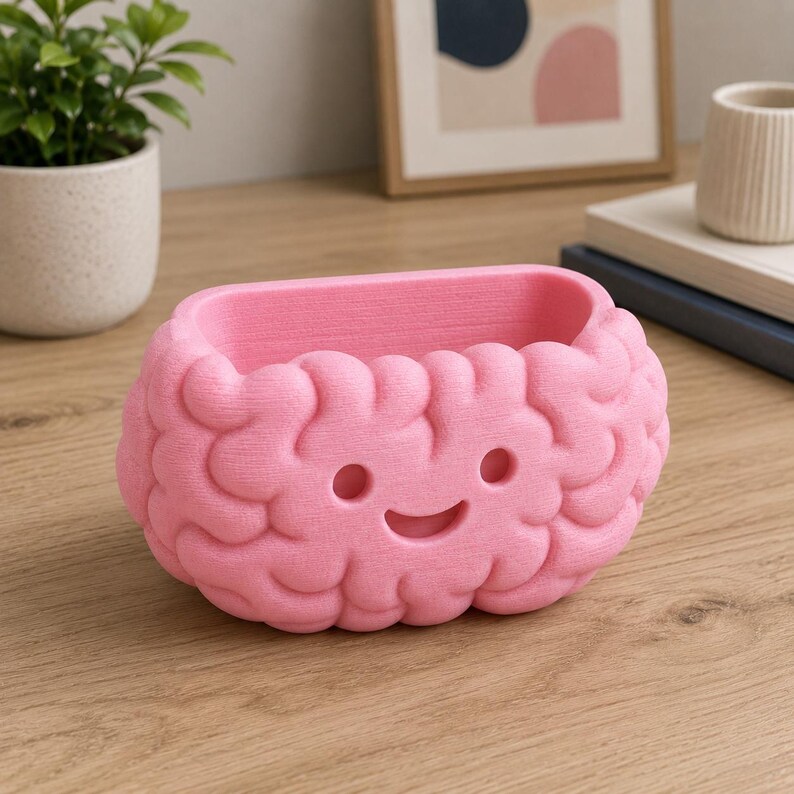 May include: A pink, brain-shaped planter with a smiling face. The planter is made of a textured material and is designed to hold small plants or other decorative items. The design is whimsical and fun.