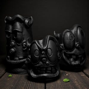 Könnte beinhalten: Drei schwarze, cartoonartige Gesichtsskulpturen mit übertriebenen Merkmalen, darunter große Augen und Münder. Die Skulpturen sind auf einer dunklen Holzoberfläche mit dunklem Hintergrund angeordnet. Die Gesichter haben ein strukturiertes Aussehen.