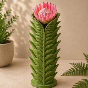 Pu&ograve; includere: Un vaso verde con un design a forma di felce, che contiene un fiore di protea rosa. Il vaso ha una forma cilindrica con motivi di foglie dettagliati. Il fiore &egrave; di un rosa vibrante con un centro strutturato, creando un contrasto sorprendente.