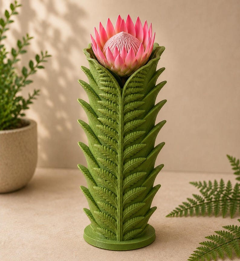 Pu&ograve; includere: Un vaso verde con un design a forma di felce, che contiene un fiore di protea rosa. Il vaso ha una forma cilindrica con motivi di foglie dettagliati. Il fiore &egrave; di un rosa vibrante con un centro strutturato, creando un contrasto sorprendente.