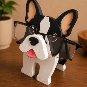 May include: A black, white, and tan wooden eyeglasses holder shaped like a French Bulldog. The dog has a black nose, pink tongue, and is holding a pair of eyeglasses. The holder is on a wooden surface.