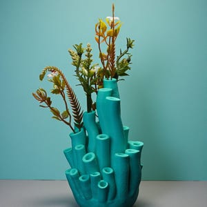 May include: A teal-colored, coral-shaped vase holds an arrangement of artificial flowers and foliage. The vase has multiple cylindrical openings of varying sizes. The flowers include white, yellow, and brown elements, creating a contrast against the vase's color.
