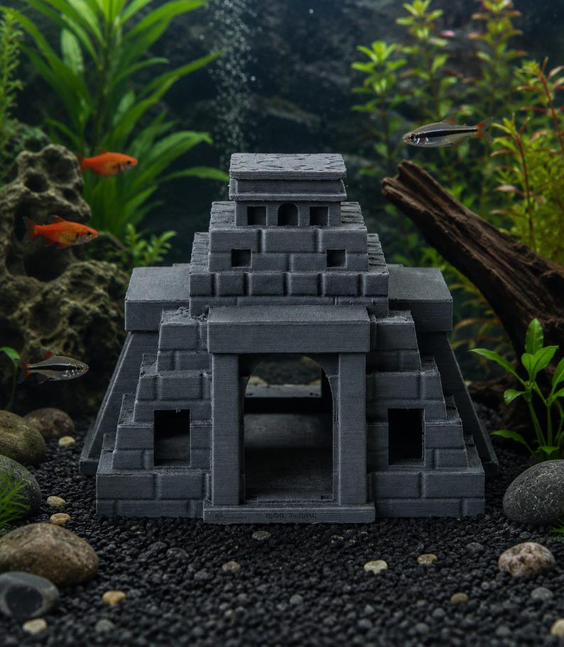 Puede incluir: Decoraci&oacute;n de acuario con forma de templo maya gris. La estructura presenta un dise&ntilde;o escalonado con aberturas rectangulares y un techo plano. La imagen muestra el templo sumergido en un acuario con peces vivos y plantas acu&aacute;ticas.