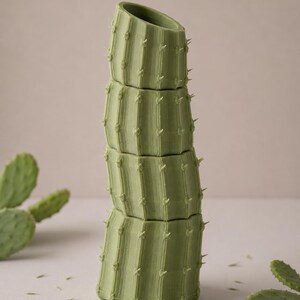 May include: A stack of four green cactus-shaped planters. Each planter section has vertical ridges and small, pointed protrusions. The top planter is angled. The background includes a few green cactus pads.
