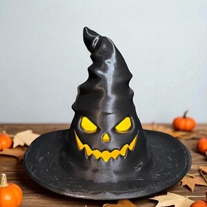 May include: A black Halloween witch's hat with a carved jack-o'-lantern face. The hat has a wide brim and a tall, pointed crown. The eyes and mouth of the jack-o'-lantern glow with a yellow light. Pumpkins and leaves surround the hat.