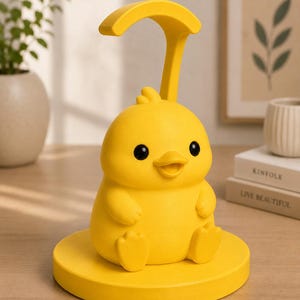 May include: A bright yellow duck-shaped desk lamp. The lamp features a rounded base, a duck body with small wings and feet, and a curved handle. The duck has black eyes and an open beak. The lamp is designed to sit on a desk or table.