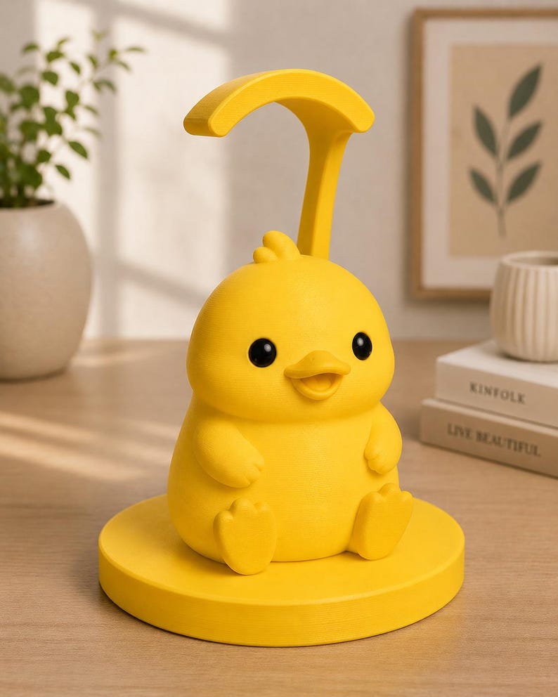 May include: A bright yellow duck-shaped desk lamp. The lamp features a rounded base, a duck body with small wings and feet, and a curved handle. The duck has black eyes and an open beak. The lamp is designed to sit on a desk or table.
