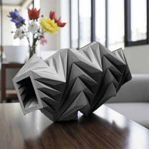 May include: A geometric, abstract sculpture in shades of gray sits on a dark wood surface. The sculpture has a complex, folded design with sharp angles and a hollow opening. A vase of colorful flowers is in the background.