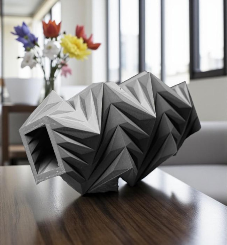 May include: A geometric, abstract sculpture in shades of gray sits on a dark wood surface. The sculpture has a complex, folded design with sharp angles and a hollow opening. A vase of colorful flowers is in the background.