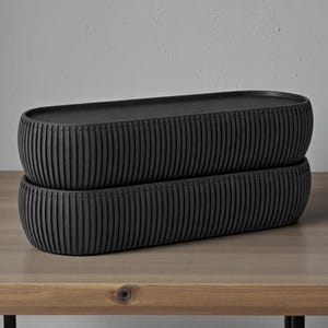 May include: Two stacked, black, oval ceramic containers with vertical ribbed detailing. The containers sit on a light brown wooden surface. The containers have a modern, minimalist design.