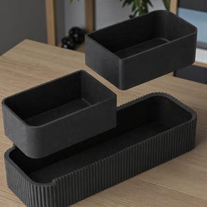 May include: Three-piece black storage containers. The set includes two rectangular boxes and a larger, grooved tray. The boxes are designed to fit inside the tray. The containers are made of a textured material and are displayed on a wooden surface.
