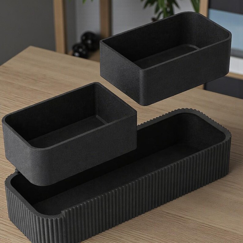 May include: Three-piece black storage containers. The set includes two rectangular boxes and a larger, grooved tray. The boxes are designed to fit inside the tray. The containers are made of a textured material and are displayed on a wooden surface.