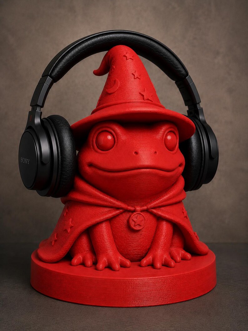 May include: A red frog-shaped headphone stand wearing a wizard hat and cape, with black headphones resting on top. The stand has a round base and is made of a solid material. The headphones are black with the Sony logo.