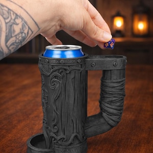 May include: A black, intricately carved beer stein with a handle, holding a blue and silver can. A person's hand is dropping a blue and purple die into the stein. The stein has a detailed, textured design, suggesting a fantasy or medieval theme.