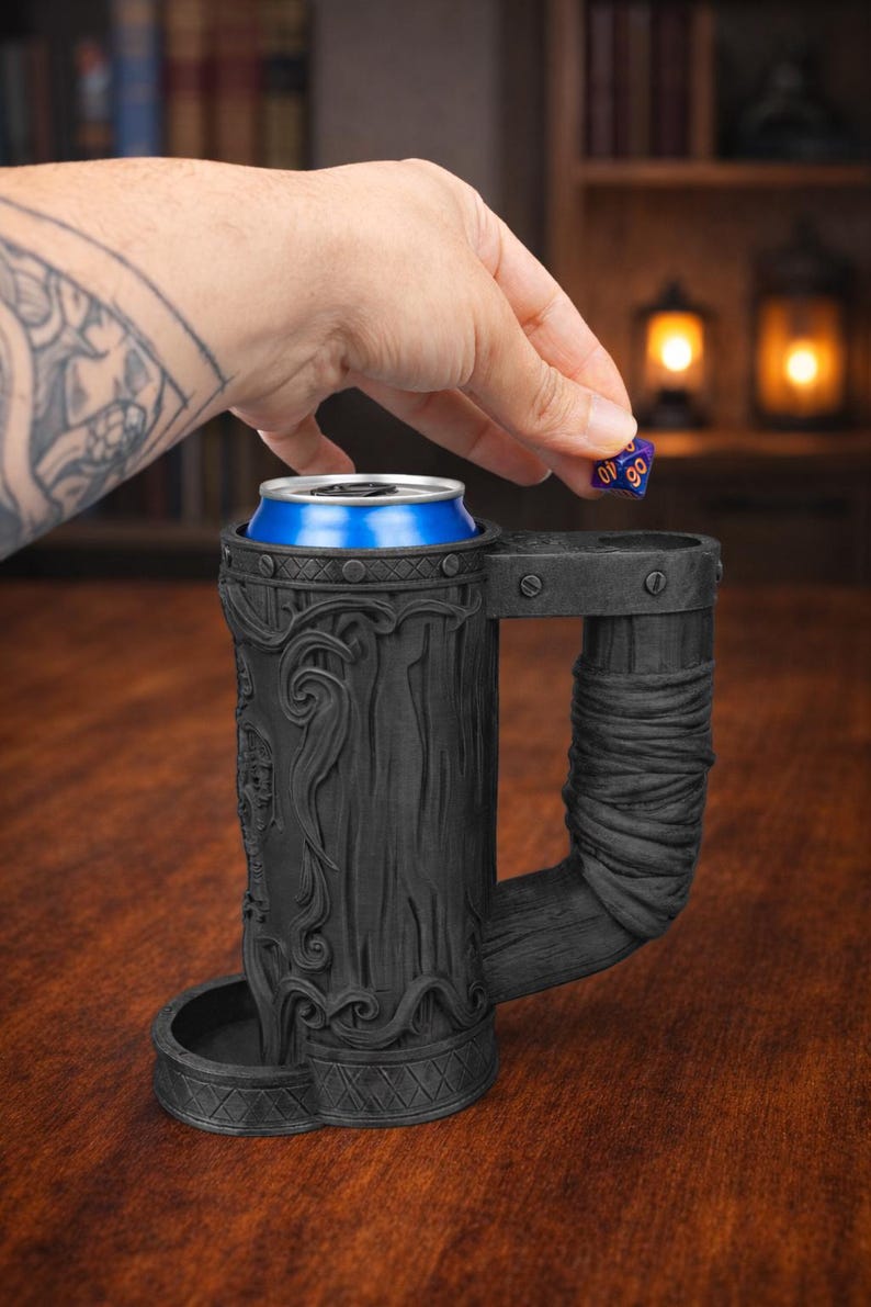 May include: A black, intricately carved beer stein with a handle, holding a blue and silver can. A person's hand is dropping a blue and purple die into the stein. The stein has a detailed, textured design, suggesting a fantasy or medieval theme.