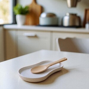 Puede incluir: Una cuchara de madera clara descansa sobre un reposacucharas de cer&aacute;mica moteada. El reposacucharas tiene una forma org&aacute;nica y curvada. El entorno de la cocina incluye una mesa blanca y un fondo borroso con utensilios de cocina.