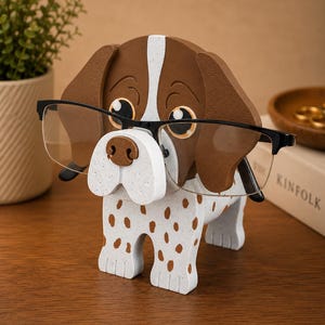 May include: A wooden dog-shaped eyeglasses holder. The holder is painted white with brown spots and a brown head with a white stripe. Black-framed glasses rest on the dog's nose. The dog is on a wooden surface.