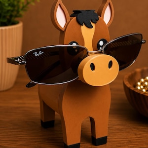 May include: A brown wooden horse-shaped eyeglass holder with a light orange nose and ears. The horse is holding a pair of Ray-Ban sunglasses with dark lenses. The holder has black hooves and a black mane.
