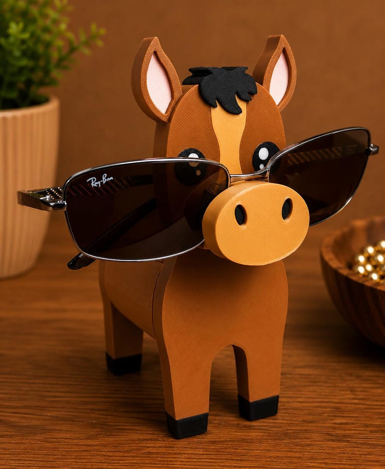 May include: A brown wooden horse-shaped eyeglass holder with a light orange nose and ears. The horse is holding a pair of Ray-Ban sunglasses with dark lenses. The holder has black hooves and a black mane.