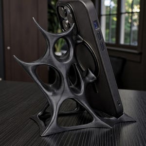 May include: A black, abstract-design phone stand with a smartphone in a black case. The stand has a unique, open structure with multiple points and curves. The phone is positioned at an angle, showcasing its camera and screen. The stand is on a dark surface.